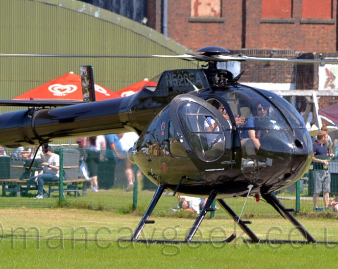 Front side view of a glossy black helicopter parked facing to the right on grass, with the motor slowly spinning and the pilots door open. The grey registration "N520EA" is on the side of the engine pods on top of the fuselage, and grey text "MD520N" is on the top of the twin tails at the end of the long, fat tail boom. Notably, there is no vertical rotor at the end of the tail boom. There are 2 people sitting in the heavily glazed cockpit, the person sitting in the right-hand seat wearing a white shirt, and using their hands to demonstrate something to the person in the left hand seat, who is wearing a blue shirt. Green grass fills the foreground, with people sitting or standing behind a green metal mesh fence in the background, and a curved, green corrugated metal hangar behind them on the left, and a large brown brick building on the right. Pretty much everyone in the photo is wearing sunglasses, so it is probably a sunny day.