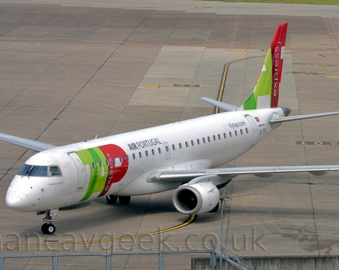 Slightly elevated side view of a twin-engined jet airliner taxiing from right to left. The plane is mostly white, with billboard-style, red and green "TAP" titles on the side of the forward fuselage, with smaller "Air Portugal " titles above the windows at the front of the passenger cabin. The tail has part of those same large red and green titles, with white "Express" titles arranged vertically in the long stroke of the red letter "P". Grey concrete apron marked with lines for planes to follow fills the rest of the frame.