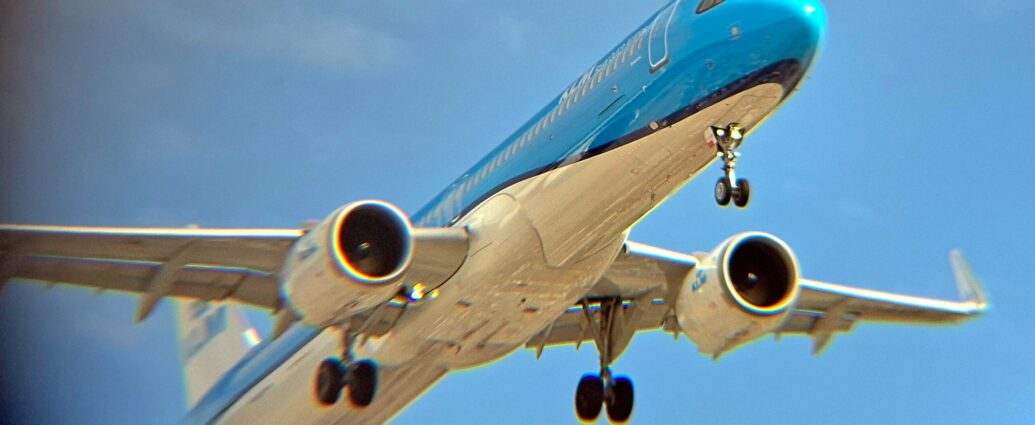 Low side view of a twin engined jet airliner flying from left to right at a very low altitude with undercarriage lowered and flaps extended from behind the wings, suggesting it is about to land. The plane is mostly a glossy light blue with a white belly, the 2 colours separated by a dark blue stripe running along the body. There are white "KLM" titles on the upper forward fuselage, with blue "KLM" titles on the sides of the white tail and white engine pods. The rear fuselage and the wing-tips are blurred and grainy, with blooms of colour along shiny surfaces and edges. Lush blue sky fills the background, with hints of cloud on the left of the frame.