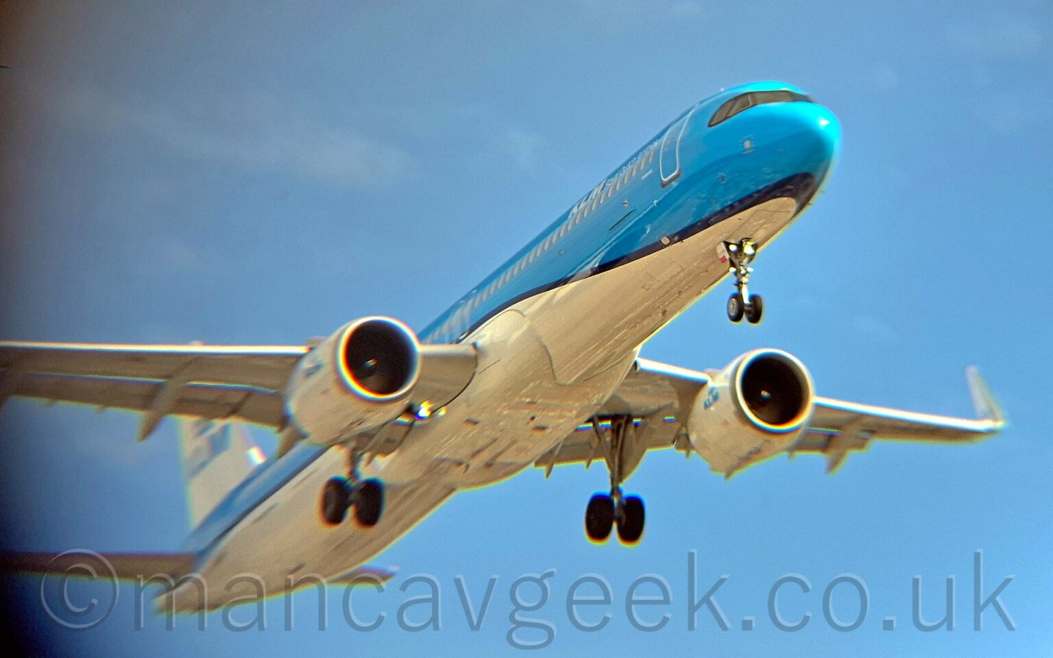 Low side view of a twin engined jet airliner flying from left to right at a very low altitude with undercarriage lowered and flaps extended from behind the wings, suggesting it is about to land. The plane is mostly a glossy light blue with a white belly, the 2 colours separated by a dark blue stripe running along the body. There are white "KLM" titles on the upper forward fuselage, with blue "KLM" titles on the sides of the white tail and white engine pods. The rear fuselage and the wing-tips are blurred and grainy, with blooms of colour along shiny surfaces and edges. Lush blue sky fills the background, with hints of cloud on the left of the frame.