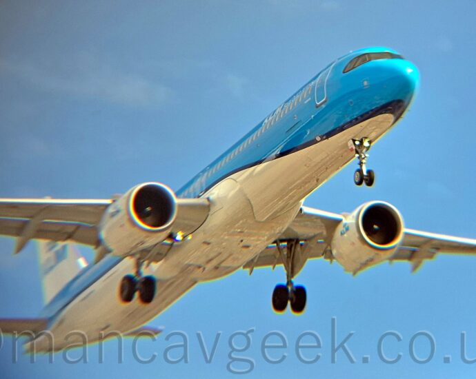 Low side view of a twin engined jet airliner flying from left to right at a very low altitude with undercarriage lowered and flaps extended from behind the wings, suggesting it is about to land. The plane is mostly a glossy light blue with a white belly, the 2 colours separated by a dark blue stripe running along the body. There are white "KLM" titles on the upper forward fuselage, with blue "KLM" titles on the sides of the white tail and white engine pods. The rear fuselage and the wing-tips are blurred and grainy, with blooms of colour along shiny surfaces and edges. Lush blue sky fills the background, with hints of cloud on the left of the frame.