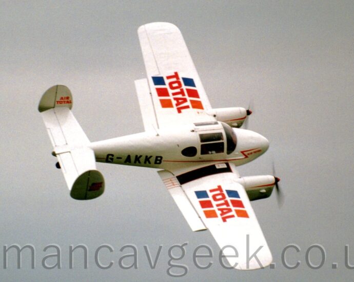 Side view of a twin engined light aircraft flying from left to right, highly banked to the right. The plane is mostly white, with a thin red stripe running along the body, overlaid with the black registration "G-AKKB" on the rear fuselage. There are red "Total" logo's on each wing, with diagonal blue, red, and orange stripes running through, and smaller red "Air Total" titles on both the inside and outside faces of the twin tails. Grey sky fills the rest of the frame.