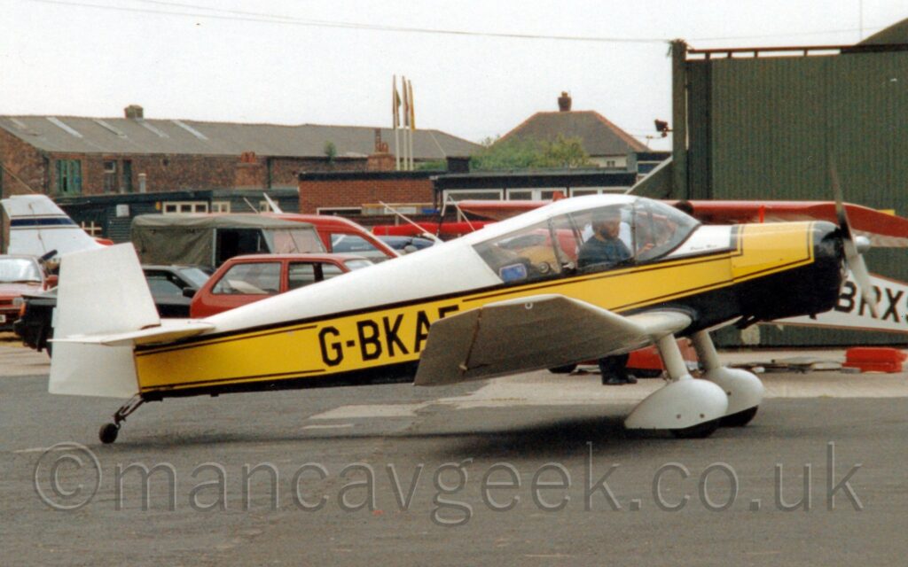 Side view of a single engined light aircraft parked on tarmac facing to the right. The plane is mostly white, with a black belly, and a thick yellow stripe running along the body, over-laid with the black registration "G-BKAE" on the rear fuselage, the last letter partially obscured by the slightly up-turned wing-tip. White spats cover the fixed main landing gear, under the wings. A man with short dark hair and beard, wearing a blue top, can be seen in the cockpit, possibly running an engine test, as the propellor on the nose looks to be slightly blurred. Black tarmac apron fills the foreground, with a green hangar on the right of the frame in the background, with cars parked behind the plane on the left, with brown brick buildings beyond that. Flat white sky fills the rest of the frame.