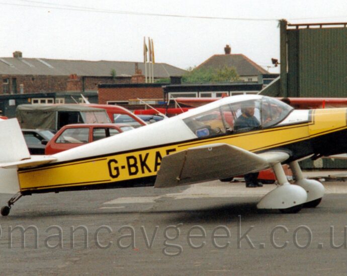 Side view of a single engined light aircraft parked on tarmac facing to the right. The plane is mostly white, with a black belly, and a thick yellow stripe running along the body, over-laid with the black registration "G-BKAE" on the rear fuselage, the last letter partially obscured by the slightly up-turned wing-tip. White spats cover the fixed main landing gear, under the wings. A man with short dark hair and beard, wearing a blue top, can be seen in the cockpit, possibly running an engine test, as the propellor on the nose looks to be slightly blurred. Black tarmac apron fills the foreground, with a green hangar on the right of the frame in the background, with cars parked behind the plane on the left, with brown brick buildings beyond that. Flat white sky fills the rest of the frame.
