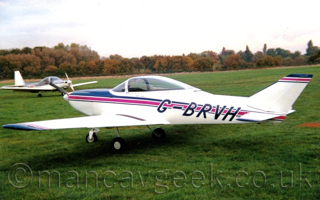Side view of a single-engined light aircraft parked on grass facing to the left. The plane is mostly white, with a large bubble canopy over the canopy, and a purple stripe running along the body, outlined in dark blue. A similar stripe adorns the top of the tail. Green grass fills the foreground, as well as most of the background, with trees in the distance. A grey and white light aircraft is parked on the left of the frame. Bright but hazy skies fill the top of the frame.