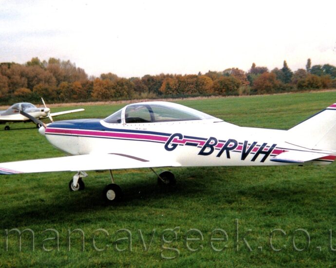 Side view of a single-engined light aircraft parked on grass facing to the left. The plane is mostly white, with a large bubble canopy over the canopy, and a purple stripe running along the body, outlined in dark blue. A similar stripe adorns the top of the tail. Green grass fills the foreground, as well as most of the background, with trees in the distance. A grey and white light aircraft is parked on the left of the frame. Bright but hazy skies fill the top of the frame.