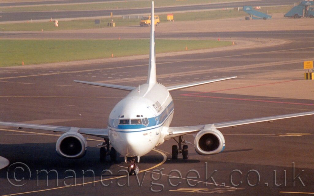 Almost head-on view of a twin engined jet airliner taxiing slightly to the left of the camera, following a curving yellow line on the black taxiway. The plane is largely white, with a blue stripe running along the body, covering the passenger cabin windows, and black "IcelandAir" titles on the upper forward fuselage. Black tarmac taxiways fill the foreground and a large chunk of the background, with yellow and red stripes marked on the surface. More black tarmac wends it's way through large islands of grass surrounded by grey concrete, with scattered yellow airport vehicles and parked blue mobile airstairs.