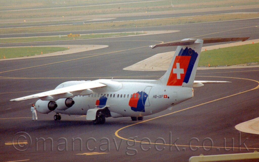 Rear side view of a high-winged, 4 engined jet airliner taxiing from right to left and away from the camera. The plane is mostly white, with a large red and blue spatchcock on the side of the rear fuselage, a smaller but similar splotch under the wings but above the open main landing gear doors, and another large one on the white tail, with the red splash replaced with a red oblong with a large white cross in the middle. The black registration "HB-IXF" is on the side of the rear fuselage, next to a small, red and white Swiss flag. Black tarmac fills the foreground, marked with yellow lines for planes to follow to their gates, with islands of green grass surrounded by grey concrete filling the background with black taxiways weaving through them.