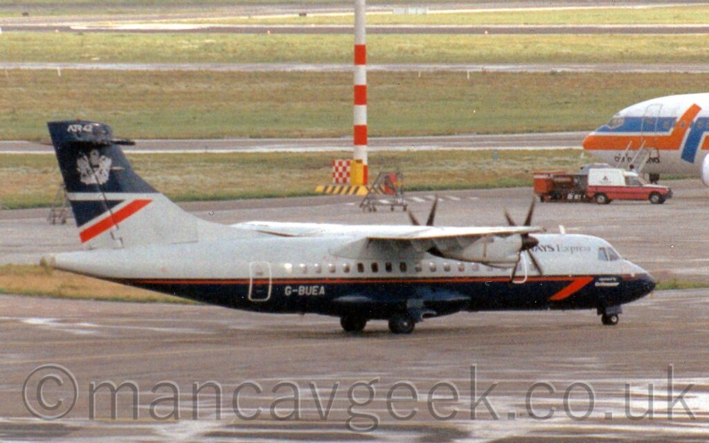 Side view of a high-winged, twin propellor engined airliner taxiing from left to right and slightly away from the camera. The plane is grey on top with a dark blue lower half, with a red pinstripe running along the body, ending in a half arrowhead below and just aft of the cockpit windows, and white text "Operated by CityFlyer Express". There is black partial text "Ways Express" on the upper forward fuselage, the start obscured by the wing, and the grey registration "G-BUEA" on the lower rear fuselage. The tail is grey at the bottom, with a dark blue and red triangle at the top rear of the grey, with a dark blue upper half with a large, grey heraldic crest towards the top, with grey text "ATR42" right at the top. Black tarmac taxiways fill the foreground, with wide strips of green grass separated by more black taxiways filling the background. The nose of a white jet airliner with thick orange and blue stripes running across the body pokes into frame from the right, with a small pink and white van parked in front.