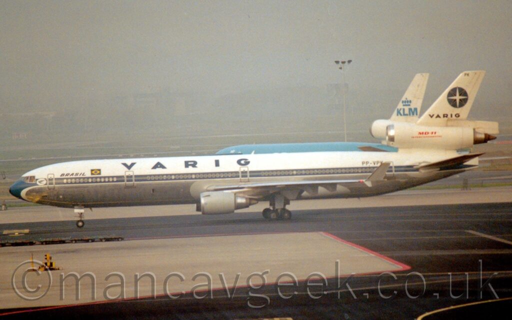 Side view of a 3 engined jet airliner taxiing from right to left. The plane is largely white, with a blue stripe running along the body, covering the passenger cabin windows, with large blue "Varig" titles on the upper forward fuselage. Additional blue text "Brazil" next to a green and yellow Brazilian flag on the forward fuselage, immediately aft of the forward cabin door. The black registration "PP-VPK" is on the upper rear fuselage. The centre engine pod runs through the tail, with large red "MD-11" titles over smaller red "Intercontinental" text. The top of the tail has black "Varig" text at the bottom, and the letters "PK" right at the top, and a large blue circle in the middle, with 4 black diagonal compass points, and 4 vertical/horizontal white points. Black taxiways fill the foreground, with a grey apron area outlined in right coming into frame from the left. A vaguely similar type of plane with a blue body and a white tail is behind this plane. Green grass and grey taxiways in the background are rapidly being swallowed up in grey mist, which fills the top of the frame.