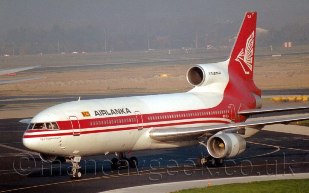 Front-side view of a 3 engined jet airliner taxiing from right to left. The plane is largely white, with a thick red stripe running along the body, covering the passenger cabin windows, and black "Air Lanka" titles on the upper forward fuselage. The red stripe sweeps up and down to fill the tail and rear fuselage. A stylised white bird with out-spread wings is on the tail. The planes centre engine intake is on the top of the fuselage, running through the bottom of the tail, and exhausting through the tail cone, with black "TriStar" text on the sides of the white intake. The white registration "4R-ULA" is on the side of the lower rear fuselage, with the black letters "ULA" on the open nose wheel doors, and white letters "ULA" on the top of the tail. Black tarmac taxiways fill most of the foreground, with a patch of green grass surrounded by grey concrete in the bottom right corner. More black taxiways surround a large, yellowing patch of grass in the background, with more grass leading up to cars on a road in the distance, with trees beyond vanishing into grey haze.