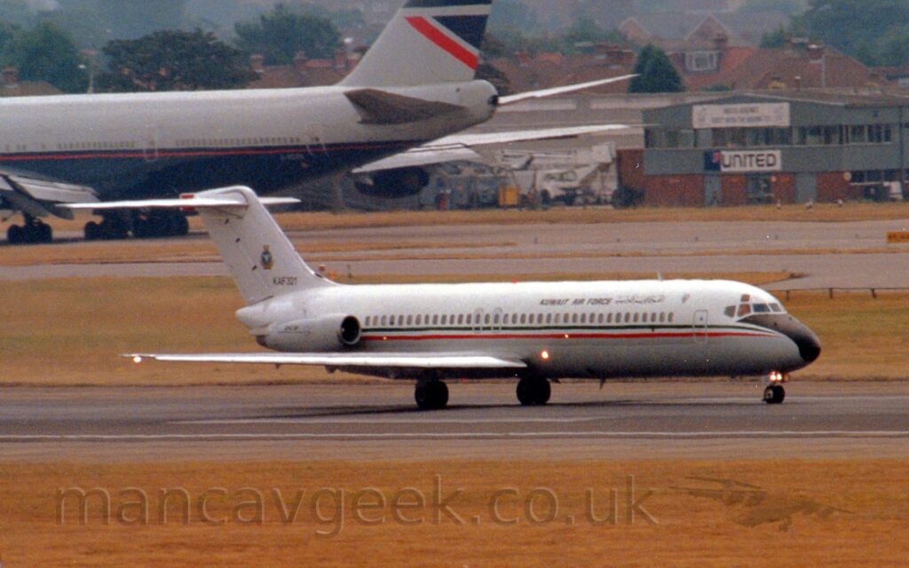 Side view of a twin engined jet airliner accelerating along a black runway, with the engines mounted on the sides of the rear fuselage. The plane is mostly white, with a grey belly, and a thin red and green stripe running along the body, covering the passenger cabin windows. There are black "Kuwait Air Force" titles on the upper forward fuselage, in an Arabic script towards the front, and in English further back. The tail is white, with the black serial "KAF321" at the base, and the emblem of the Kuwait Airforce in the middle. Yellow grass lines the runway in the foreground and part of the background, with grey taxiways beyond that4, with a very large, 4 engined jet airliner taxiing out of frame in the top left corner and a grey and red office building in the top right. Brown brick houses and trees beyond that lead off into the distance.