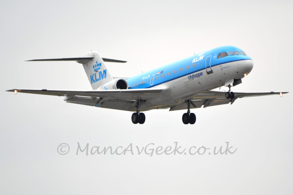 Low side view of a twin engined jet airliner with the engines mounted on the sides of the rear fuselage, lying from left to right at a low altitude, with undercarriage lowered and flaps extended from the rear of the wings, suggesting it is about to land. The plane is blue on the top half and white at the bottom, the 2 halves separated by a thin dark blue stripe. There are white "KLM" titles under a stylised white crown on the upper forward fuselage, and the white registration "PH-KZB" on the upper rear, just aft of small EU and Dutch flags. Small, blue text "Air France KLM" is on the lower forward fuselage under the cockpit windows, slightly forward of the main door, with larger "Ctiyhopper" titles just aft. There are large blue "KLM" titles under a stylised blue crown on the tail, replicated in a smaller form on the sides of the engine pods, partially obscured by the wings. Flat grey sky fills the rest of the frame.