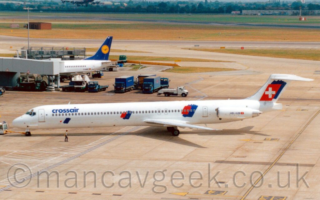 Side view of a white, twin engined jet airliner with the engines mounted on the sides of the rear fuselage, facing to the left but being pushed back to the right from it's gate by an almost-out-of-frame white truck attached to the nosewheel by a long yellow pole. There are blue "CrossAir" titles on the upper forward fuselage, with red and blue splotches on the sides of the fuselage just afore and aft of the wing, repeated on the tail overlaid with a white cross. The registration "HB-IUM" is on the side of the engine pods in black. Concrete apron fills the foreground, with taxiways and a runway separated by grass in the background. A grey airbridge stretches in to frame from the left, attached to a white, twin engined jet airliner with a blue and yellow tail.
