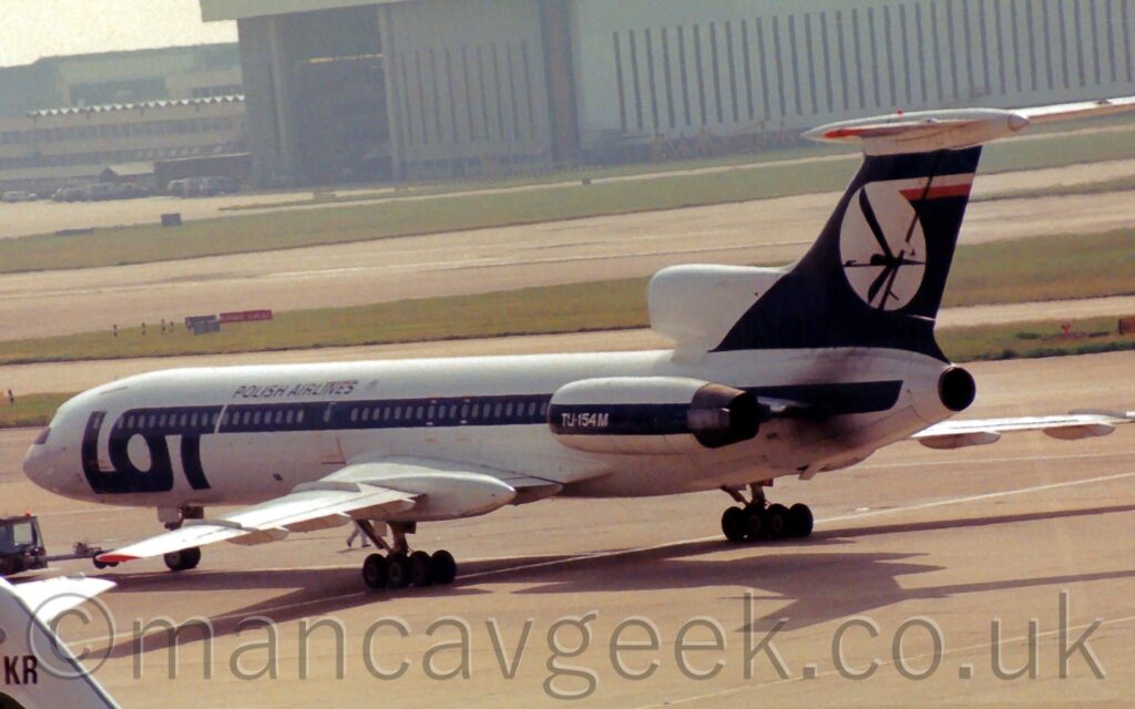 Side view of a 3 engined jet airliner taxiing from right to left. The plane is mostly white, with large dark blue "Lot" titles on the forward fuselage, the cross-bar on the "T" stretching all the way back along the cabin windows to the tail-mounted engine, as well as along the engine pods mounted on the sides of the rear fuselage. There are small, blue "Polish Airlines" on the upper forward fuselage, and white text "TU-154M" on the blue stripe on the side engine pod. The tail is dark blue, with a white circle containing a stylised blue flying bird, as well as a white and red stripe at the top of the tail. Grey concrete taxiway fills the foreground, with large grassed areas lining more taxiways in the background, and a large grey hangar in the distance.