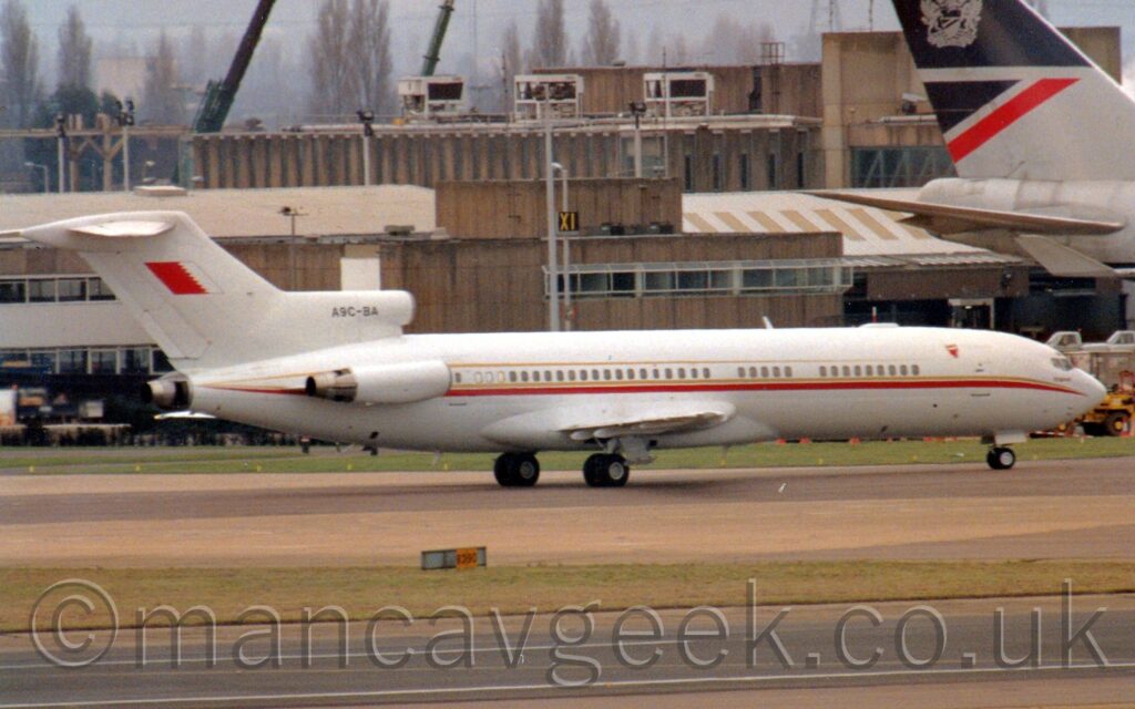 Side view of a white 3 engined jet airliner with the engines mounted on the rear fuselage, taxiing from left to right. There is a gold and red stripe running along the body just below the passenger cabin windows, with a second gold stripe just above the windows, and a reversed Bahrain flag (Large red field next to a large white field, separated by a jagged edge - normally displayed the other way around) on the tail. The registration "A9C-BA"is on the centre engine inlet in black. A black runway stretches across the bottom of the frame, separated from this plane by a strip of grass. A brown building stretches across the background, with the grey rear fuselage and tail of a very large jet airliner is on the right of the frame. Leafless trees stand out against grey sky beyond the building.