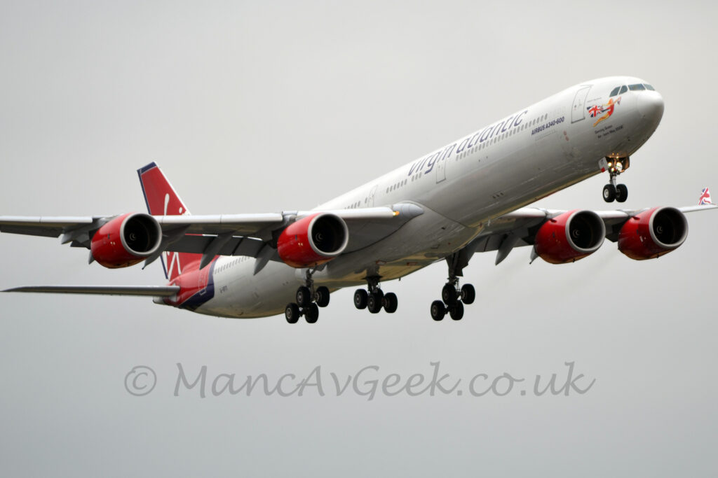 Side view of a 4 engined jet airliner flying from left to right at a low altitude, with flaps extended from behind the wings and undercarriage lowered, suggesting it is about to land. The plane is largely white, with purple "Virgin Atlantic" titles on the upper forward fuselage, and a pair of red engine pods under each wing. The tail is red, outlined in dark blue, with seemingly random white white lines. There is an image of a flying woman wearing a red mini-dress and trailing a waving British flag from the back of her dress and outstretched arms. There are 2 lines of purple text under the cockpit windows - "Dancing Queen", and "Air - Born May 2006", and additional text "Airbus A340-600" below the forward passenger cabin windows. The registration "G-VFIT" is on the lower rear fuselage, with the last 2 letters "IT" on the open nose gear doors. Flat grey sky fills the background.