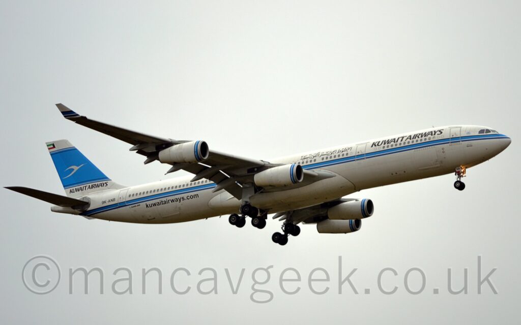 Low side view of a 4 engined jet airliner flying from left to right at a low altitude, with undercarriage lowered and flaps extended from the rear of the wings, suggesting it is about to land. The plane is white, with a blue and black stripe running along the body, under the passenger cabin windows. There are black "Kuwait Airways" titles on the upper forward fuselage, in English at the front and in an Arabic script a bit further back. The black web address "KuwaitAirways[dot]com" on the lower rear fuselage, and the black registration "9K-ANB" on the upper rear. The tail is white, with black "Kuwait Airways" titles at the bottom and the Kuwaiti flag at the top, and a wide blue stripe filling the rest, with a stylised flying white bird overlaid on top. The black and blue stripe along the fuselage is replicated as rings around the front of the engine pods under the wings. Flat grey sky fills the rest of the frame.