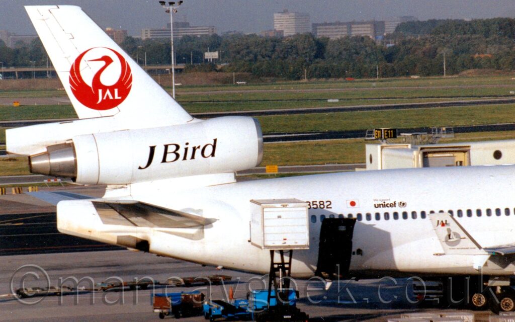 Side view of the white rear fuselage and tail of a 3 engined jet airliner parked on it's stand facing to the right. The partial registration "8582" is on the upper rear fuselage, next to a small Japanese flag and small Unicef logo, the start of the registration obscured by the bed of a catering truck that is raised up to the rear door. A cargo door is hinged upwards is the lower rear fuselage, next to the catering truck. The centre engine pod runs through the tail, and has large black text "J Bird" on the side. The top of the tail has a stylised image of a red bird with it's wings stretched out, overlaid with the white letters "JAL". These letters are repeated in black on the up-turned wingtips, over a grey-scale image of a bird flying across a grey circle. Grey apron fills the bottom of the image, with large grassed areas criss-crossed by black taxiways in the background, and trees and tall buildings in the distance, under grey skies.