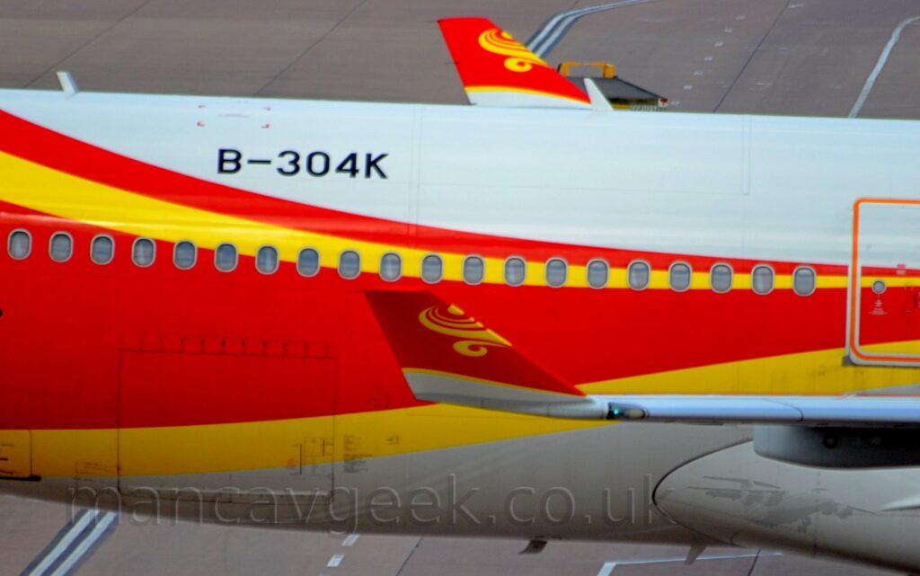 Close-up side view of the central fuselage and up-turned wingtips of a jet airliner taxiing from left to right. The plane is white on top, with a grey belly, and a thick red stripe running from front to rear, getting wider and moving down towards the lower rear fuselage and up into the tail, as well as a yellow stripe along the lower part of the red stripe. The wing-tips are similar, mostly red with yellow swirls and a white base. Grey concrete with painted white lines apron fills the background.