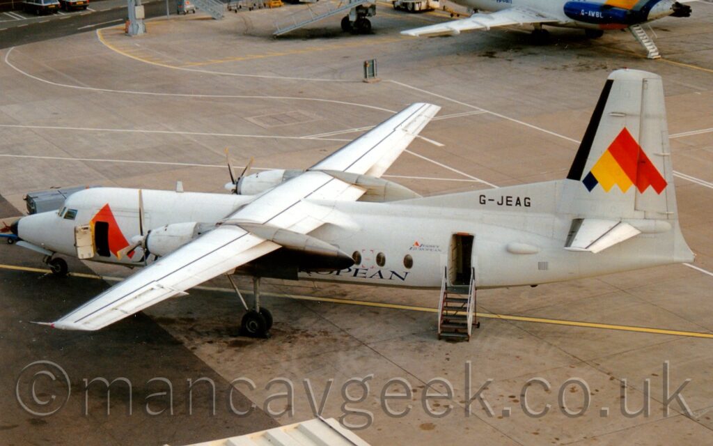 Elevated side view of a high-winged, twin propellor-engined airliner parked facing to the left. The plane is mostly white, with a large, upward pointing triangle nade of diagonal black, orange, red, and yellow stripes and a serrated bottom edge, a design replicated in a smaller form over the open forward cabin door, and the black registration "G-JEAG" on a white fillet where the front of the tail joins the top of the fuselage. A small set of mobile airstairs is parked against the open rear cabin door, which has small "Jersey European" titles and an even smaller version of the triangular logo just in front of it. Squares of grey concrete apron marked with yellow and white lines fill the rest of the frame, with the left wing and rear fuselage of a white, twin engined jet airliner peeking into shot at the top.