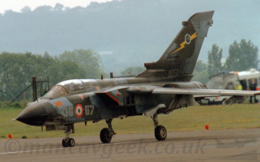 Side view of a twin engined military jet fighter aircraft parked facing to the left. The plane is covered in a grey and green-brown camouflage scheme, with the white numbers "36" and"57" either side of a red, white, and green roundel under the 2 seat cockpit, and a yellow lightning flash overlaid with a black circle containing an image of a bird on the tail. Black runway fills the foreground, with grass in the background on the far side of the runway leading up to trees at the airfields edge. Tree-covered hills rise in the distance, slowly vanishing into the murky grey skies.