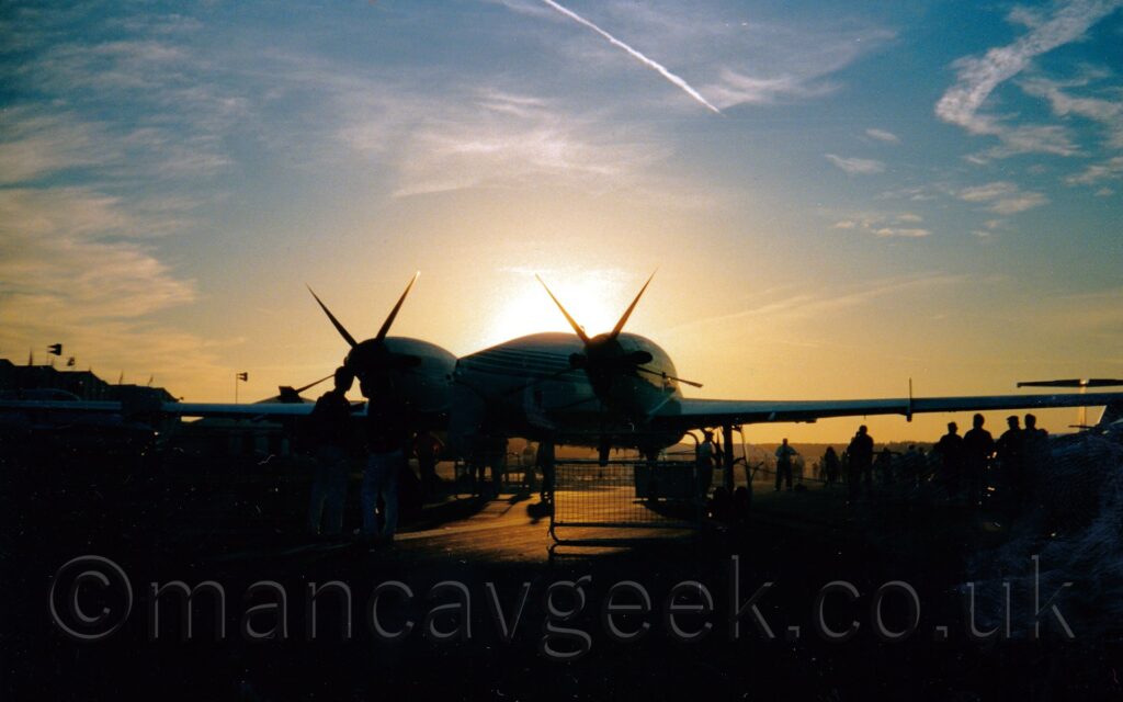 Rear view of a twin pusher-propellor-engined BizProp, with the engines mounted on the rear of the wing, parked facing away from the camera. The plane is mostly white, with thin blue stripes running along the body, just above the wings. The sun is setting directly behind the planes body, leaving a lot of the foreground in shadow, with only occasional bits illuminated. Several people can be seen standing around in the foreground and background, including 2 people standing in front of the left engine. Bright blue sky and wisps of cloud fill the top of the frame, with a yellow bloom around the top of the plane.