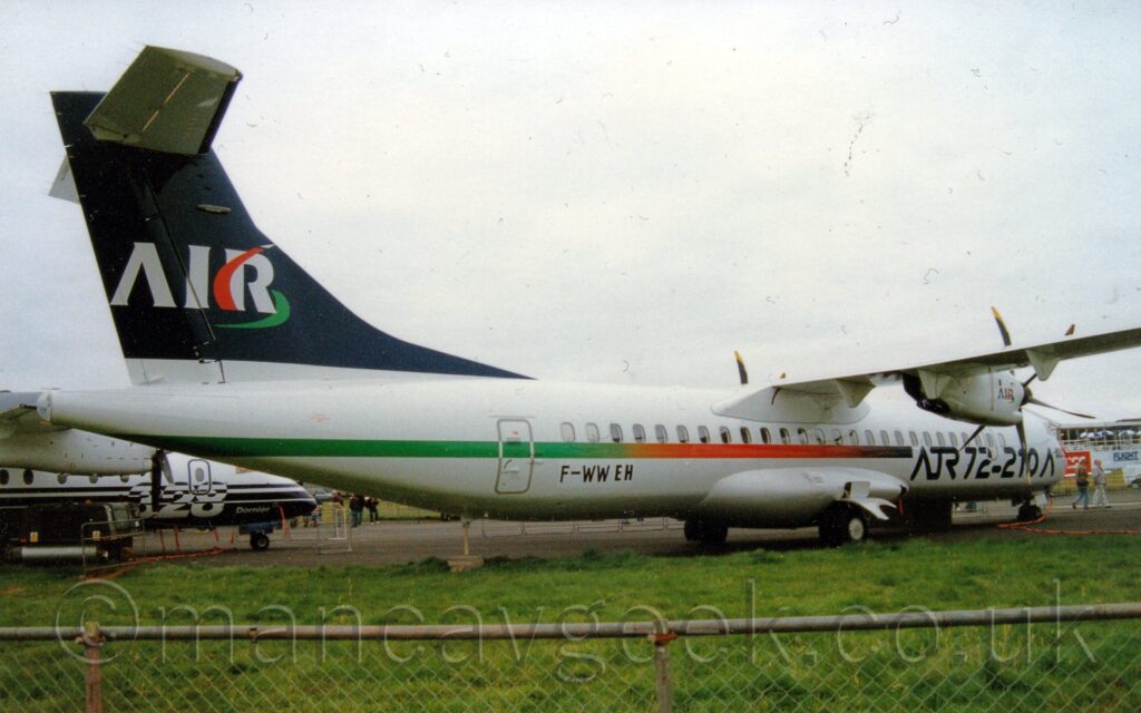 Side view of a high-winged, twin propellor-engined airliner parked facing to the right and slightly away from the camera, behind a metal fence. The plane is largely white, with a multi-coloured stripe running along the body, dark blue towards the front, then orange in the middle, and then from yellow to green towards the rear, above the black registration "F-WWEH" on the lower rear fuselage. There are large, black, "ATR 72-210" titles on the lower forward fuselage. Green grass fills the foreground, behind a chainlink metal fence. A smaller, blue and white high-winged airliner can be seen in the background under the tail of this plane on the left, with a white open-sided building on the right. Flat white sky fills the rest of the frame.