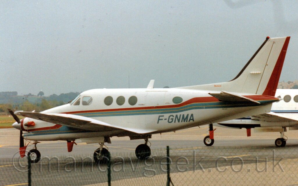 Side view of a twin propellor-engined BizProp parked facing to the left on grey tarmac. The plane is mostly a creamy-white, with a red, grey, and yellow stripe running along the body, as well as along the sides of the engine pods in front of the wings. The grey registration "F-GNMA" is on the side of the lower rear fuselage. A black chain-link fence stretches along the bottom of the frame. Another white BizProp can be seen in the background on the right, with trees and fields on the left, under flat grey sky.