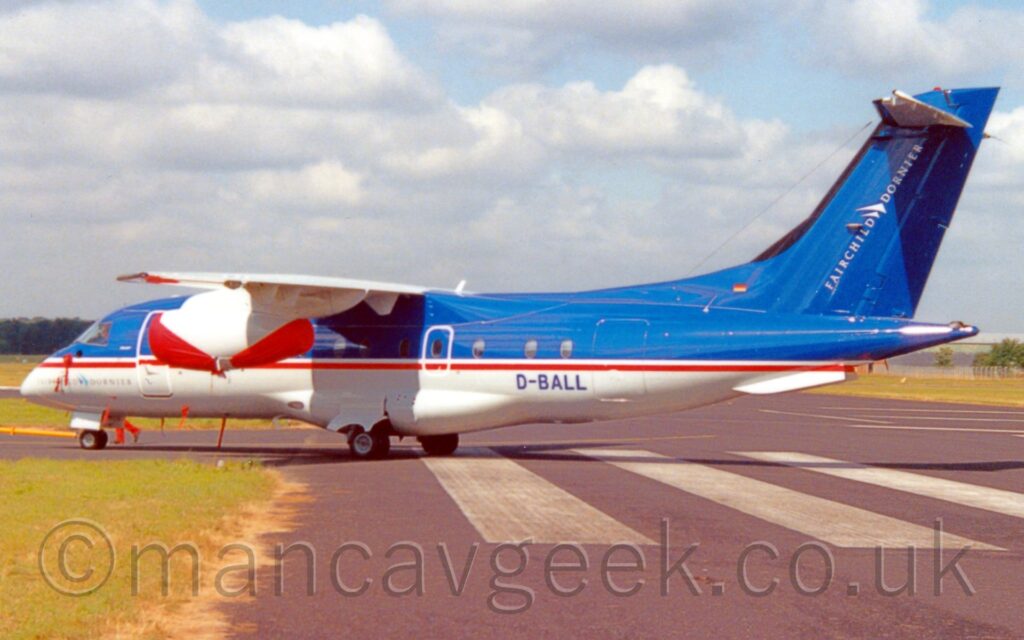 Side view of a high-winged, twin engined jet airliner parked facing to the left on a grey taxiway. The plane has a blue top half and white bottom, with a thin white and red stripe under the passenger cabin windows separating the two sections. There are diagonal "Fairchild Dornier" titles on the rear of the blue tail, and the blue registration "D-BALL" on the lower rear fuselage. A grey taxiway fills most of the foreground, with a block of green grass in the bottom left corner, Fluffs of white and grey cloud with patches of blue peeking through fill the top of the frame.
