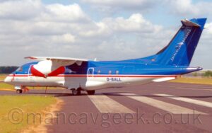 Side view of a high-winged, twin engined jet airliner parked facing to the left on a grey taxiway. The plane has a blue top half and white bottom, with a thin white and red stripe under the passenger cabin windows separating the two sections. There are diagonal "Fairchild Dornier" titles on the rear of the blue tail, and the blue registration "D-BALL" on the lower rear fuselage. A grey taxiway fills most of the foreground, with a block of green grass in the bottom left corner, Fluffs of white and grey cloud with patches of blue peeking through fill the top of the frame.