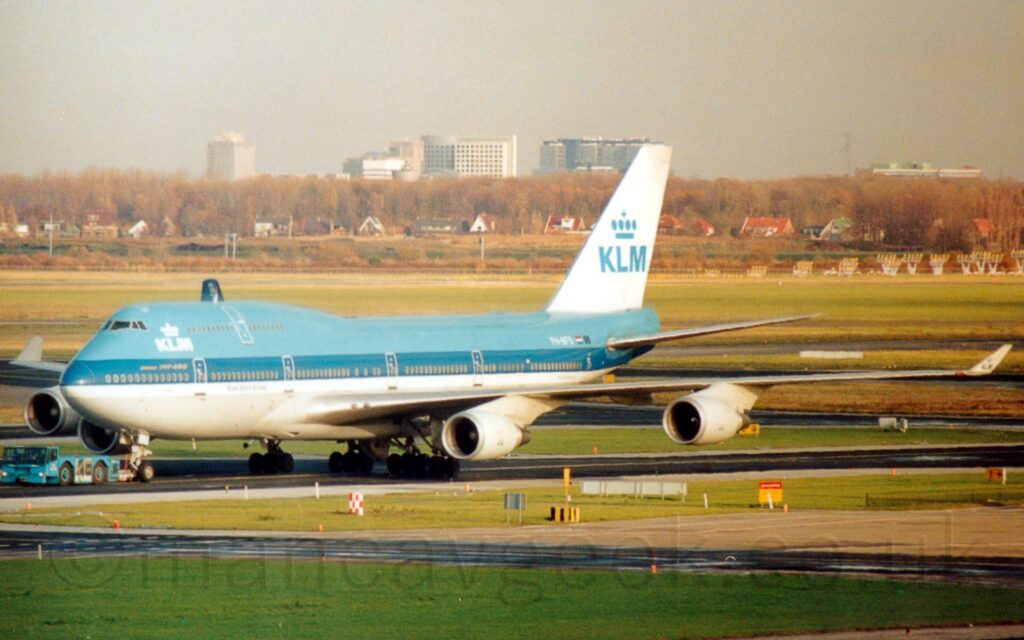 Side view of a very large, 4 engined jet airliner being towed from right to left to it's gate by a low-slung blue tug attached to the nose-wheel by a long white pole. The plane is light blue on top, with a thick dark blue stripe covering the lower passenger deck windows, , with a slightly thinner white stripe below that, and a bare metal belly. There are white "KLM" titles on the upper forward fuselage, between the cockpit windows and the upper deck windows, under a stylised white crown. This same logo appears in a larger form in light blue on the white tail. Green grass divided by a black taxiway fills the foreground, with more, larger areas of grass with taxiways winding their way through fills much of the background, with houses nestled in trees in the distance, under flat grey skies.