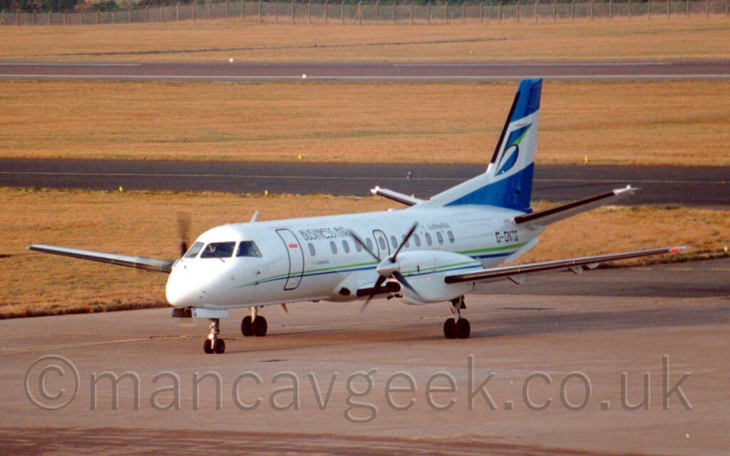 Front side view of a twin propellor-engined airliner taxiing from right to left along a grey taxiway. The plane is mostly white, with a thin green and blue stripe running along the body, with black "Business Air" titles on the upper forward fuselage, and smaller "Lufthansa" titles on the upper rear. A similar green and blue stripe runs along the sides of the engine pods mounted in front of the wings. The black registration "G-GNTG" is on the rear fuselage, below the dark blue tail. A diagonal white band fills the middle of the tail. With a highly stylised lower-case letter "b" with a black outline and seemingly random blue, green, and white triangles. Grey concrete taxiway fills the foreground, with yellowing grass and another black taxiway in the background leading up to a black runway in the distance, with more yellowing grass and a chainlink fence beyond that.