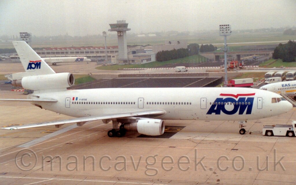 Side view of an almost all-white, 3 engined jet airliner being towed from left to right by a low-slung white tug attached to the nose-wheel. There are large, blue, billboard-style "AOM" titles on the forward fuselage, with stylised red flying bird that resembles an almost straight line over the top, a logo repeated in a smaller form on the tail. There is a smaller, white jet airliner with the same logo on it's forward fuselage in the background on the left, parked in front of a white building with multiple red slanted roofs. Next to that building, towards the centre of the image is a squat, grey building with a tall grey tower poking up from corner, with a heavily glazed top level and several clock faces suspended underneath. Grass leads off into the distance, slowly vanishing into murky grey haze.