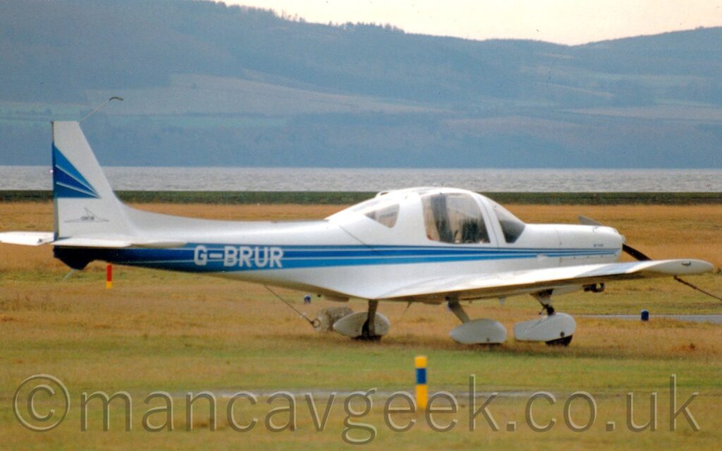 Side view of a single-engined light aircraft parked facing to the right on grass, with plastic weights attached to the wings to hold it down. The plane is white, with 3 thin blue stripes running back along the body from the nose, getting wider as they move rearwards, overlaid with the white registration "G-BRUR" on the sides of the rear fuselage. Three similar stipes appear diagonally on the tail. Green grass fills the foreground and most of the background, with a wide river in the distance, and tree-covered hills beyond that, under bright but hazy sky.