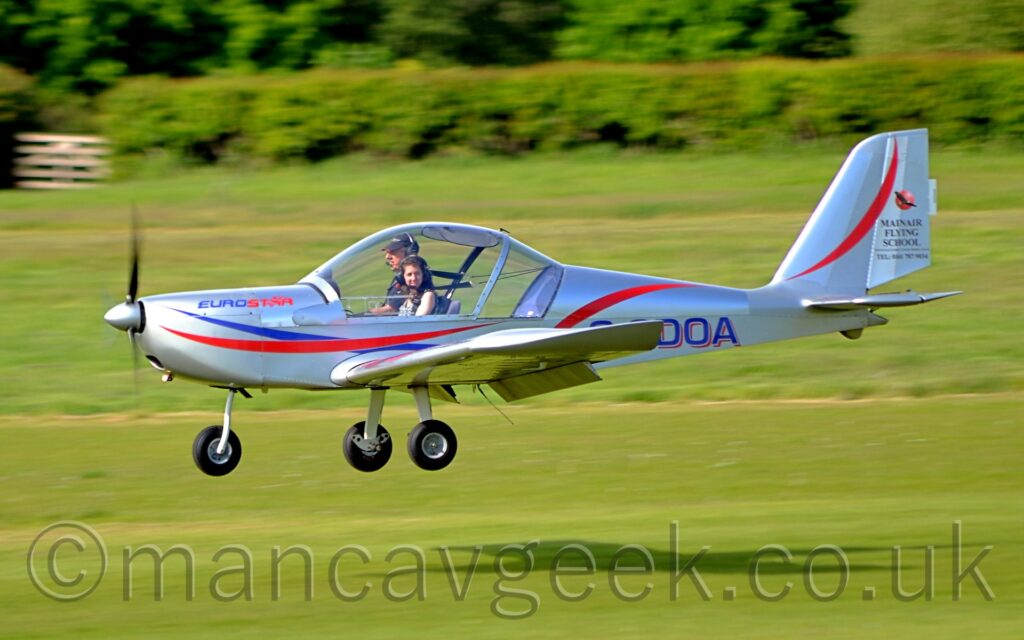 Side view of a single-engined light aircraft flying from right to left at a very low altitude, just 3 or 4 metres above a grass runway, with flaps extended from the rear of the wings, which are straight and level, suggesting it is just about to land. The plane is a silvery grey, with red and blue stripes running along the body, sweeping up and over the wings, the red stripe then going over the top of the body, and along the tail. The blue and red text "EuroStar" is on the top of the engine cowling, right in front of the cockpit. The blue registration "G-CDOA" is on the sides of the rear fuselage, the first couple of letters partially obscured by the wing. A large bubble canopy covers the cockpit, and it's two occupants, a tall man wearing a black cap and black t-shirt in the right-hand seat, and a shorter woman with long dark hair in the left-hand seat. He is looking straight ahead, while she is looking off to her left. The plane appears to be getting ever closer to it's shadow on the ground. Green grass fills the background, leading up to trees and bushes in the distance.