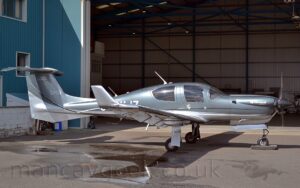 Side view of a very sleek, single-engined light aircraft parked on concrete apron facing to the right, that looks as if it is moving fast even when it is standing still. The plane is mostly a silvery grey with a white belly, and the white registration "G-CMJZ" on the side of the rear fuselage, partially obstructed by the right wing. Additional white text "DA50" is on the tail, and "Diamond Aircraft" on the engine cowling on the nose. In the foreground, a large wet patch with puddle is on the ground on the left, although the rest of the ground is bone dry. The plane is casting a strong shadow on the ground, with sunlight coming from just left of overhead. The planes nose-wheel is resting on what looks like a black plastic box, but is actually a very small, remotely controlled tug. In the background, a large blue hangar with white trim fills the frame, it's doors it's doors wide open, with a couple of residents visible inside.