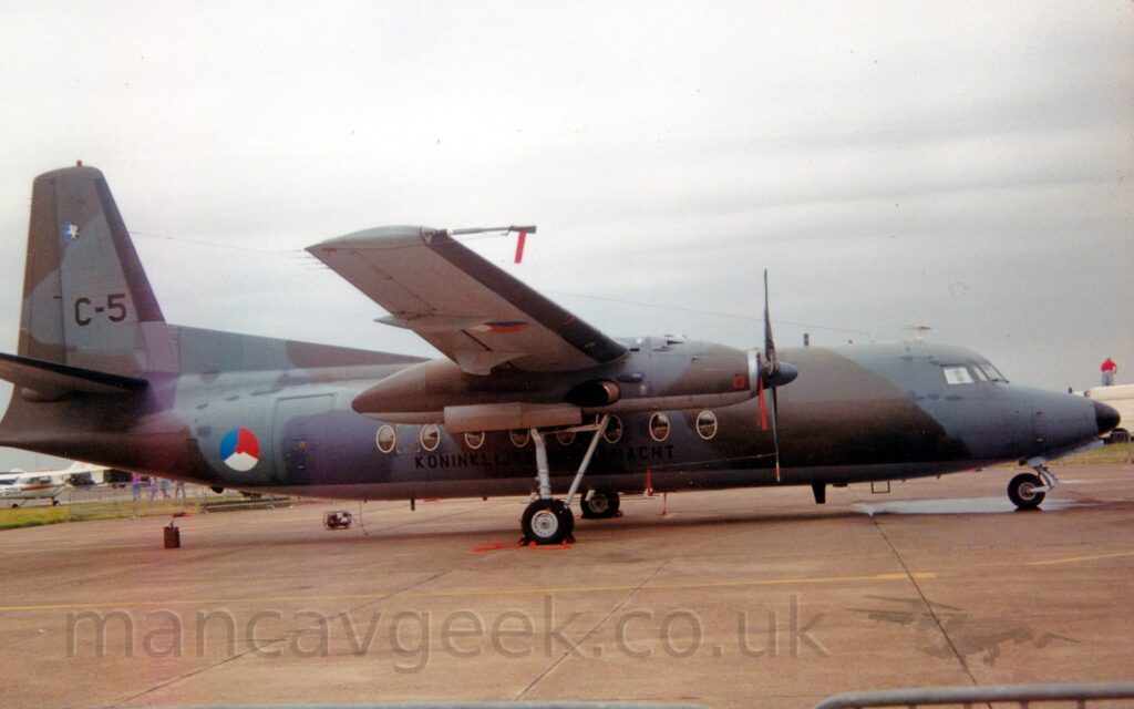 Side view of a high-winged, twin propellor-engined military transport aircraft parked facing to the right on grey concrete apron. The plane is covered in a dark grey and olive green camouflage scheme, with a light grey belly, with black text "Koninklijke Luchtmacht" (English translation - Royal Dutch Air Force) on the lower fuselage, below the cabin windows. A 3 part roundel, white on the bottom, blue on the top left, and red on the top right, with a red centre, is on the side of the rear fuselage, and repeated under the outer panels of the right wing. The large, black serial "C-5" is on the tail. Grey concrete apron fills the foreground, with flat grey sky filling the rest of the frame.