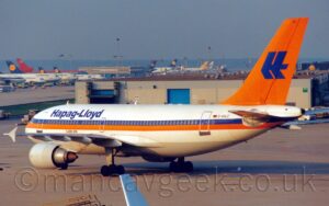 Rear side view of a twin engined jet airliner taxiing from right to left. The plane is mostly white, with a blue and orange stripe running along the body, the blue bit covering the passenger cabin windows. There are blue "Hapag-Lloyd" titles on the upper forward fuselage, and the black registration "D-AHLC" next to a small German flag on the upper rear fuselage. The tail is bright orange, with a highly stylised upper-case letter "H" in the middle. A wide brown building with a large blue door stands in the middle of the frame in the background, with rows of jet airliners lined up either side of another brown building in the distance. Grey blue sky fills the top if the image.