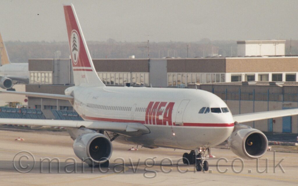 Side view of a twin engined jet airliner taxiing from left to right, following a yellow line along a grey taxiway. The plane has a white top half and a grey belly, with a red stripe running along the body, separating the 2 halves of the fuselage. There are large, red "MEA" titles on the forward fuselage, and the black registration "PH-AHE" on the upper rear. The base of the tail is white, with the top being red, with a white circle in the middle containing a stylised image of a green tree. A sandy brown building stretches across the background from the right, with a white airliner #with a yellow tail behind that on the left of the frame, under grey sky.