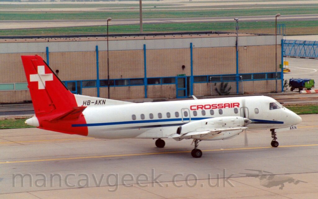 Side view of a twin propellor-engined airliner, taxiing from left to right along a grey taxiway. The plane is mostly white, with a blue and grey stripe running along the body from under the nose, and red "CrossAir" titles on the upper forward fuselage. The black registration "HB-AKN" is on a white fillet at the front edge of the tail where it joins the top of the fuselage. The tail is red, wrapping around the rear fuselage, with a large white cross in the middle. A sandy brown building with a grey roof fills most of the background from the left, with grass and runways in the distance.