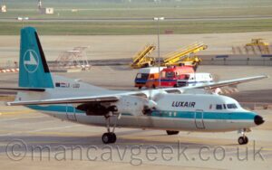 Side view of a high-winged, twin propellor-engined airliner taxiing from left to right, following a yellow line marked on grey concrete apron. The plane is mostly white, with a thick, light blue stripe running along the body, covering the passenger cabin windows, and a grey belly. There are black "LuxAir" titles on the upper forward fuselage, and the black registration "LX-LGD" next to small EU and Luxembourg flags on a white fillet at the front of the tail. The tail itself is the same light blue, with the white outline of a circle containing the stylised image of a white flying bird in the middle. Grey concrete apron fills the foreground, with even more apron in the background, with several multi-coloured airport vehicles parked behind the plane, along with yellow portable baggage loading belts, with acres of grass in the distance.