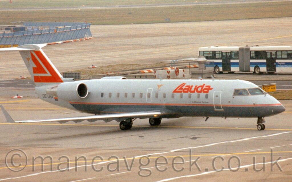 Three-quarter front side view of a twin engined jet airliner with the engines mounted on the rear sides of the fuselage, taxiing from left to right across a taxi way covered in white road markings. The plane is light grey on top and dark grey on it's lower half, with a thin red stripe running along the body, separating the 2 sections. There are red 'Lauda" titles on the upper forward fuselage, the letter "L" being made up of a couple of nestled chevrons pointing to the bottom left. A smaller, inverted, version of those chevrons fill the light grey tail. Grey apron fills the foreground and most of the background, with a white bendy bus with a blue stripe along the body is parked on the right of the frame, and a blue mesh metal fence separates out another area on the left. Brown grass and a black runway run across the top of the frame.
