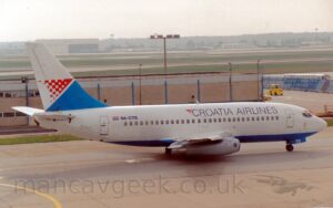 Side view of a twin engined jet airliner taxiing from left to right, following a yellow line along a grey taxiway. The plane is largely white, with a blue belly, and black "Croatia Airlines" titles along the upper forward fuselage., with the black registration "9A-CTB" on the upper rear, next to a small Croatian flag. There is a blue triangle at the base of the white tail, under a red triangle made of a white and red checkerboard pattern. Grey taxiway fills the foreground, while a sandy brown building stretches across the background from the left. Green grass and black runways lead up to a cream coloured hangar in the distance, with trees and hills beyond that, under hazy grey sky.