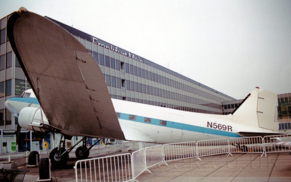 Side view of a World War 2-era, twin propellor-engined airliner on display facing to the left on the roof of an airport terminal building. The plane is mostly white, with a blue stripe running along the body, covering the square passenger cabin windows, and the black registration "N569R" on the upper rear fuselage. White metal temporary fencing snakes it's way around the body, with the wing on the left of the frame hanging out over the fencing. The heavily glazed frontage of an even taller, grey and black section of the building fills most of the background, with silvery "Frankfurt Main" titles on the top, under flat white sky.