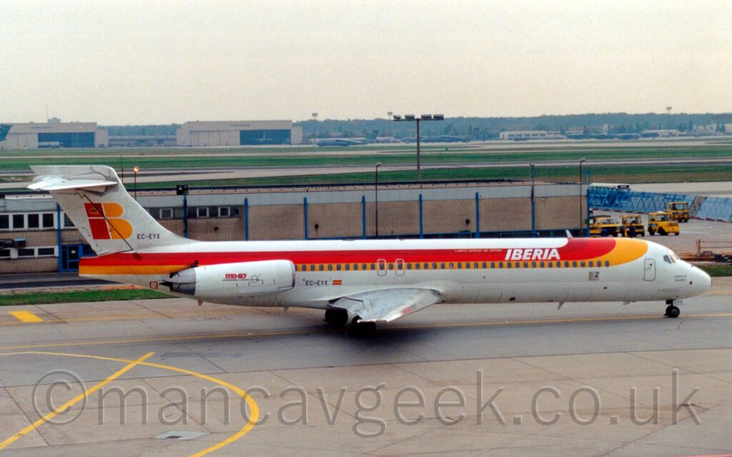 Side view of a twin engined jet airliner with the engines mounted on the sides of the rear fuselage, taxiing from left to right, following a yellow line marked on a grey taxiway. The plane is largely white, with a yellow and red stripe coming down from the top of the forward fuselage then running straight to the rear fuselage, covering the passenger cabin windows. There are white "Iberia" titles on the red part of the stripe on the upper forward fuselage, and red and yellow "MD-87" text on the side of the white engine pods on the sides of the rear fuselage. The black registration "EC-EYX" is on the base of the tail, below a red lower-case letter "I" and a yellow upper-case letter "B". The registration is repeated on the lower rear fuselage, just ahead of the engines, and just aft of a small Spanish flag. Grey concrete apron fills the foreground, with a brown and grey building stretching across most of the background from the left, with more apron on the right. Wide strips of green grass and runways lead up to cream coloured hangars and green military transport aircraft in the distance, under bright but hazy sky.