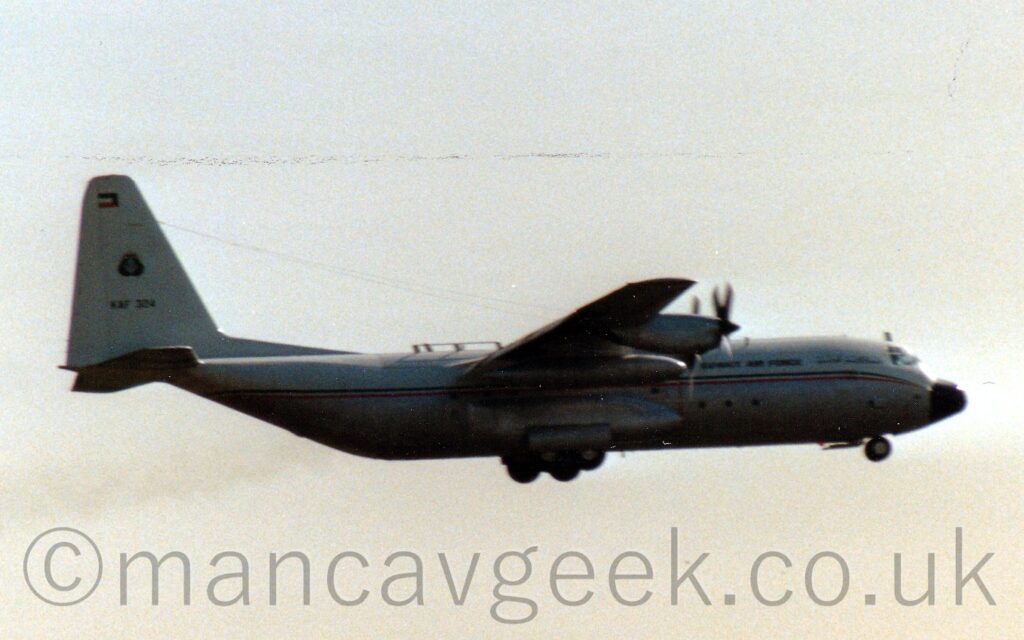 Side view of a high-winged, 4 propeller engined military cargo plane flying from left to right at a very low altitude, with undercarriage lowered, and flaps extended from the rear of the wings, suggesting it is about to land. The plane has a white top half, with a grey lower half, separated by a thin, green, white, and red stripe running along the body, and "Kuwait Air Force" titles on the upper forward fuselage. The black serial "KAF324" is on the base of the tail, below the Kuwait Air Force insignia. Bright but hazy sky fills the background.