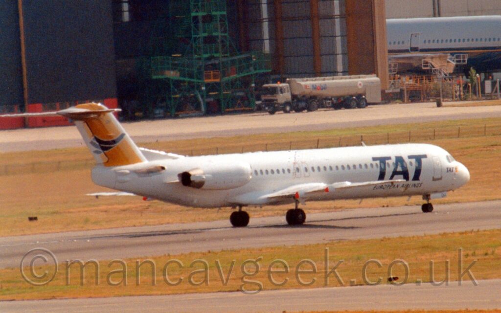 Side view of a twin engined jet airliner with the engines mounted on the sides of the rear fuselage rolling along a black runway. The plane is mostly white, with large, blue, billboard-style "TAT" titles on the forward fuselage. The tail is yellow, with a blue, highly stylised flying bird in the middle. Green grass and a grey taxiway line the side of the runway in the foreground, with more grass in the background leading up to a brown hangar with open black doors in the distance.