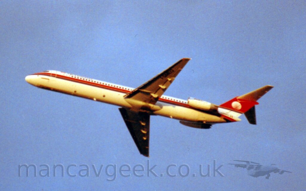Side view of a twin engined jet airliner with the engines mounted on the sides of the rear fuselage flying from right to left at a low altitude, with undercarriage retracted, flaps extended from the rear of the wings, and the nose raised sharply, suggesting it has just taken off. The plane is mostly white, with a black and red stripe running along the body underneath the passenger cabin windows, and black "Meridiana" titles on the upper forward fuselage. The black registration "I-SMEU" is under the outer panels of the left wing, as well as on the tail cone, next to a small Italian flag. The tail is white at the bottom, the rest being red with a white circular logo in the middle. Vivid blue sky fills the rest of the frame.