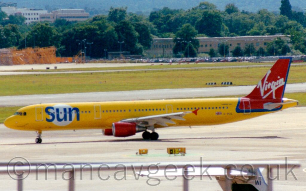Side view of a bright yellow, twin engined jet airliner taxiing from right to left across a large expanse of grey apron. There is a large white circle on the forward fuselage, overlaid with the blue word "Sun", and red engines under the wings, as well as red vertical wingtips. The tail and the top of the rear fuselage is red, outlined in blue, with whiter, seemingly hand-written "Virgin" titles in the middle. Grey apron fills the foreground, with a grey metal railing on top of a building right at the bottom of the frame. A grey runway lined on both sides by grass is in the background, with trees beyond that leading off into the distance.